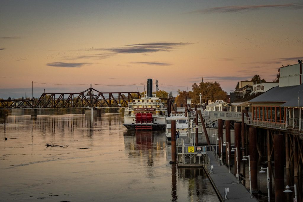 A scenic view of a classic steamboat by the river at sunset, with a historic bridge in the background.